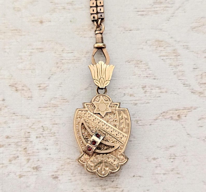 May include: A gold-toned pendant with a Star of David and intricate engraved details. The pendant features a shield shape with a decorative floral design at the bottom and a small band with three red gemstones.