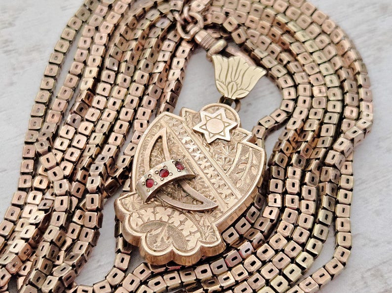 May include: Gold-toned pendant necklace with a shield-shaped pendant and a Star of David. The pendant has a ring with red gemstones. The necklace chain is made of small, square links.