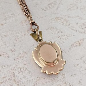 May include: A vintage gold-toned pendant necklace. The pendant has an oval center and a decorative shield-like frame. The chain is a delicate, linked design. The piece is set against a light-colored, textured surface.