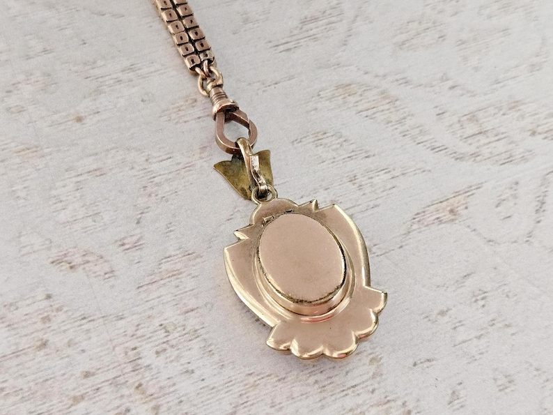 May include: A vintage gold-toned pendant necklace. The pendant has an oval center and a decorative shield-like frame. The chain is a delicate, linked design. The piece is set against a light-colored, textured surface.
