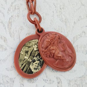 May include: A vintage-style pendant necklace in a reddish-brown hue. The pendant features an oval locket with a black and white photo and a detailed cameo profile. The necklace is suspended from a matching chain.