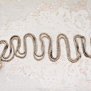 May include: A vintage gold-toned necklace with a decorative pendant. The chain is composed of small, square links arranged in a wavy pattern. The pendant features an ornate design with intricate details. The necklace rests on a textured, off-white surface.