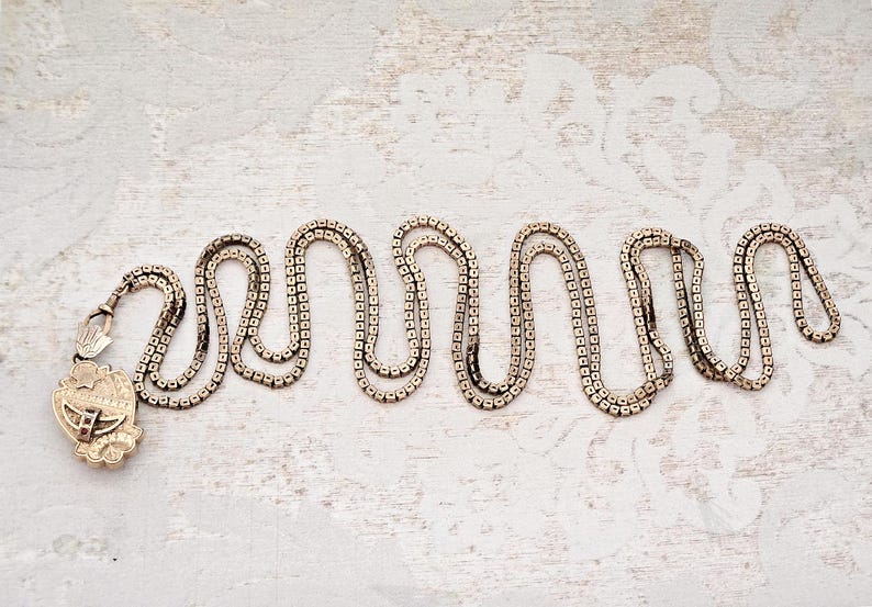 May include: A vintage gold-toned necklace with a decorative pendant. The chain is composed of small, square links arranged in a wavy pattern. The pendant features an ornate design with intricate details. The necklace rests on a textured, off-white surface.
