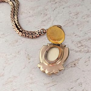 May include: A vintage gold-toned locket necklace with an open, shield-shaped pendant. The pendant reveals a cream-colored interior and a gold-colored lid. The chain is a matching gold color.