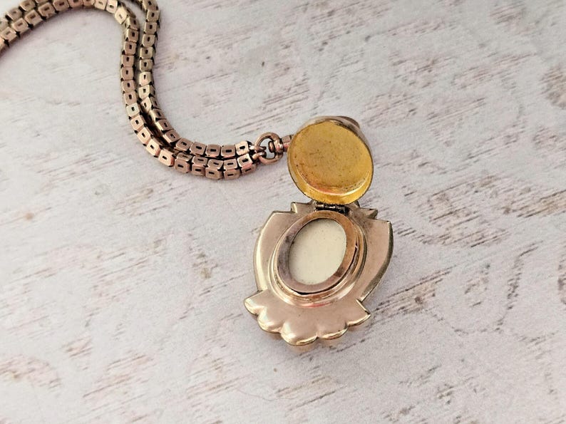 May include: A vintage gold-toned locket necklace with an open, shield-shaped pendant. The pendant reveals a cream-colored interior and a gold-colored lid. The chain is a matching gold color.