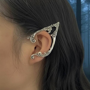 May include: Silver ear cuff with an intricate, swirling pattern. The cuff curves around the ear, with a pointed end and detailed scrollwork. The jewellery piece is worn on a person's ear, with dark hair in the background.