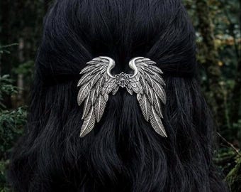 May include: A silver-toned metal hair accessory shaped like a pair of detailed wings. The wings feature intricate feather textures and are positioned horizontally, meeting at a central point with a small circular emblem in the middle.