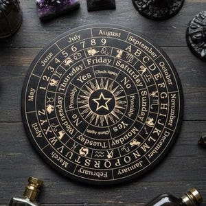 Puede incluir: Un tablero de adivinación negro y dorado con símbolos astrológicos, meses, días y el alfabeto. El tablero presenta un diseño de estrella central y las palabras "Sí", "No" y "Posiblemente". Otros objetos rodean el tablero.