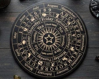May include: A circular black board featuring gold-colored engravings of the alphabet, months, days of the week, numbers one through nine, zodiac symbols, and the words Yes, No, Possibly, and Check Again, centered with a star design.