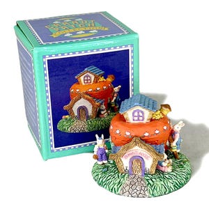 May include: A Carrot Patch Collection figurine featuring a whimsical bunny village with miniature houses atop orange carrot-shaped structures. The houses have blue roofs and are surrounded by green grass and small bunny figures. The box is teal with the collection name.