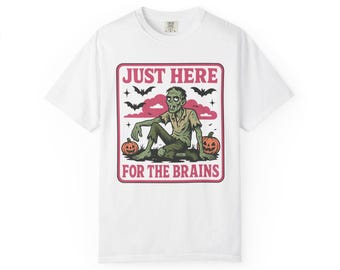 May include: White t-shirt featuring a zombie illustration with the text "JUST HERE FOR THE BRAINS". The design includes a zombie, bats, and pumpkins, perfect for Halloween. The shirt is made of a soft, comfortable material.