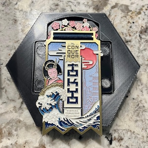 Puede incluir: Medalla con temática japonesa, con una geisha, un sol naciente y una ola estilizada. La medalla está en un marco hexagonal e incluye el texto "The Conqueror" y caracteres japoneses. Diseñada para un desafío virtual.