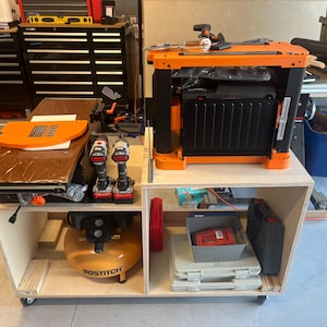 May include: A wooden workbench with two shelves. The top shelf has an orange and black planer and the bottom shelf has an air compressor, a toolbox, and other tools. The workbench is on wheels.