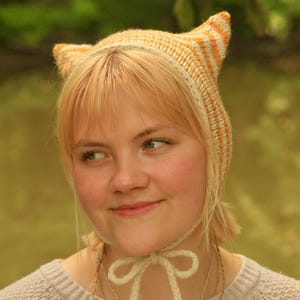 Puede incluir: Un gorro tejido a mano con dos orejas puntiagudas en forma de gato en la parte superior. El accesorio presenta un patrón de rayas en tonos crema y naranja suave. Incluye lazos largos de lana color crema a juego para atar bajo la barbilla.