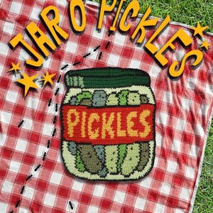 Könnte beinhalten: Ein gehäkelter Wandbehang in Form eines mit grünen Gewürzgurken gefüllten Glases, mit einem dunkelgrünen Deckel und einem roten Etikett mit dem gelben Schriftzug PICKLES. Der Text JAR O' PICKLES steht darüber, umgeben von kleinen schwarzen Ameisen.