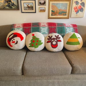 May include: Four round, off-white crocheted Christmas cushions featuring a snowman, a Christmas tree, a reindeer, and a mountain. They are arranged on a neutral-toned sofa, with a red and green patterned blanket in the background.