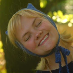 May include: A blue knitted bonnet featuring two pointed cat-like ears at the top and long, thin knitted ties that hang down from the chin area. The bonnet is crafted from a textured, heathered blue yarn and fits snugly around the head.