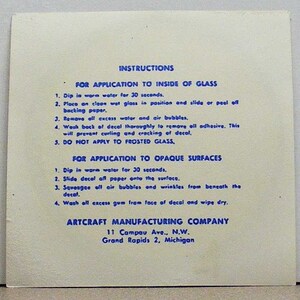 May include: A square off-white instruction sheet with blue text detailing application steps for decals on glass and opaque surfaces. The text includes instructions for water immersion, positioning, and smoothing, ending with the Artcraft Manufacturing Company address.