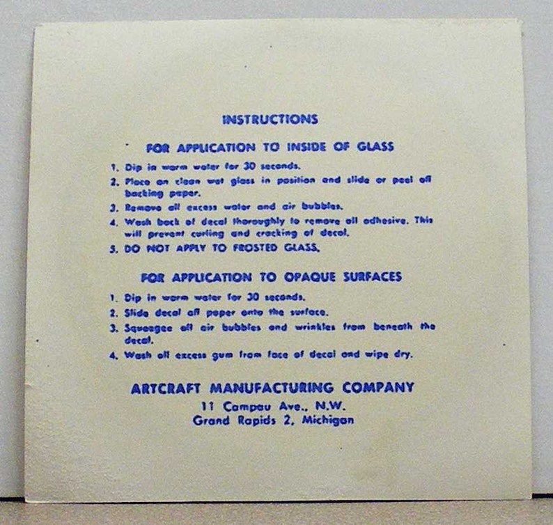 May include: A square off-white instruction sheet with blue text detailing application steps for decals on glass and opaque surfaces. The text includes instructions for water immersion, positioning, and smoothing, ending with the Artcraft Manufacturing Company address.