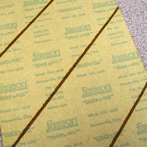May include: A rectangular sheet of tan backing paper with repeating green text that reads Fasson Crack 'n Peel, Send on Line, Painesville, Ohio, and patent numbers 3,006,793 and 3,005,957. Two dark brown diagonal lines cross the paper surface.