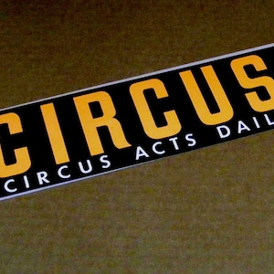 May include: A rectangular sticker with a black background and yellow lettering that reads CIRCUS H. Below the main text, smaller white lettering reads CIRCUS ACTS DAILY. The sticker is oriented diagonally across the frame with a white border.