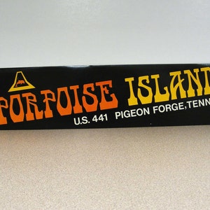 May include: A rectangular black sign with orange and yellow text reading PORPOISE ISLAND. Above the text is a yellow triangle icon containing a small orange dolphin. Below the text, white lettering reads U.S. 441 PIGEON FORGE, TENN.