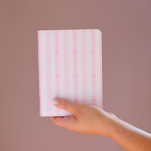 May include: A pink and white striped notebook with pink bow accents is held in a hand. The notebook has a vertical striped pattern with pink bows and dotted lines. The cover is a light pink color.