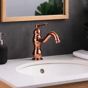 May include: A copper-colored bathroom faucet with a curved spout and a lever handle. The faucet has a classic design and is mounted on a white countertop. A black soap dispenser and a wooden-framed mirror are visible in the background.