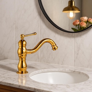 Könnte beinhalten: Ein goldfarbener Badezimmerhahn mit gebogenem Auslauf und Hebelgriff. Der Wasserhahn ist auf einer weißen Marmorarbeitsplatte über einem weißen ovalen Waschbecken montiert. Ein schwarz gerahmter Spiegel und eine Leuchte spiegeln sich im Spiegel.