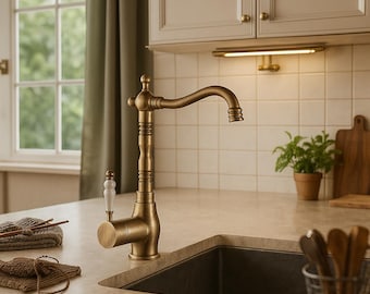 Puede incluir: Un grifo de cocina de color bronce con un caño curvo y una palanca con mango blanco. El grifo está instalado en una encimera de color claro, con un fregadero oscuro y una ventana en el fondo. El estilo es vintage.
