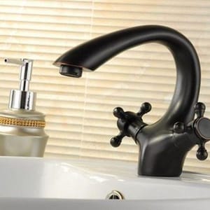 May include: A matte black bathroom faucet with a curved spout and two cross-style handles, positioned next to a silver-toned soap dispenser with a gold-colored band, both resting on a white ceramic sink basin with a visible drain hole.