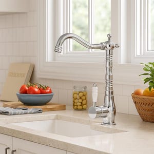 May include: A polished chrome kitchen faucet with a curved spout and decorative detailing. The faucet features a white handle and is set against a white tiled backsplash and a white sink. The image also shows a cookbook, tomatoes, and a bowl.