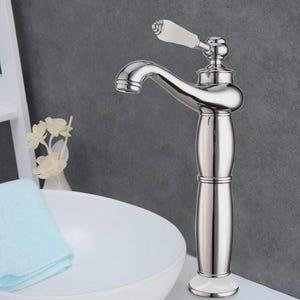Puede incluir: Un grifo de baño cromado con un mango de porcelana blanca. El grifo tiene un caño curvo y una base decorativa. El grifo está montado sobre una superficie blanca, con un lavabo blanco y una toalla azul claro en el fondo.