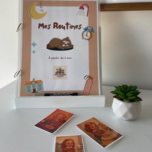Op de afbeelding: Een witte displaystandaard met een map met de tekst "Mes Routines" en illustraties van een maan, luiaard en wekker. Daaronder drie geïllustreerde kaarten met een kind. Rechts een kleine pot met vetplanten.
