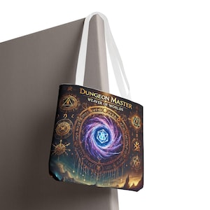 Puede incluir: Una bolsa de tela blanca con un diseño negro y dorado. El diseño presenta una galaxia giratoria con un dado azul y blanco en el centro. El texto "Dungeon Master Weaver of Worlds" está impreso en dorado en la parte superior de la bolsa.