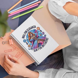 May include: A white spiral notebook featuring a colourful illustration of a woman in a blue and pink dress holding a dice. The text "Roll for Eleganza. Slay for Initiative!" is printed on the notebook.