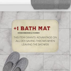 May include: A rectangular bath mat with rounded corners. The mat has the text "+1 BATH MAT" in red, with additional text below. Two gray knitted slippers are in the lower left corner.