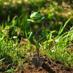 Puede incluir: Una figurita de rana verde sosteniendo una pequeña pala, de pie en un lecho de hierba verde y flores blancas.