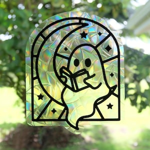 May include: Holographic sticker depicting a cartoon ghost reading a book within a stained-glass window design. The window features a crescent moon and star details. The sticker has a black outline and an iridescent finish, suitable for windows or smooth surfaces.