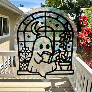 May include: A holographic window cling with a black outline design of a ghost reading a book by a window. The window features a moon, stars, and potted plants. The cling has a rainbow effect.