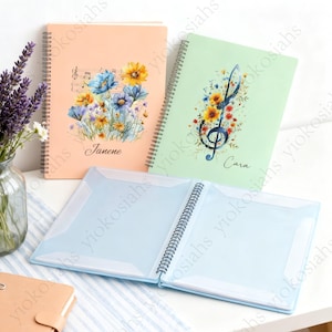 以下が含まれることがあります： 花柄のデザインが施された、3冊のリング綴じノート。1冊は、青、黄色、紫の花と「Janene」という名前が入ったピーチ色。もう1冊は、音符と花、そして「Cara」という名前が入った淡い緑色。水色のフォトアルバムが開いています。