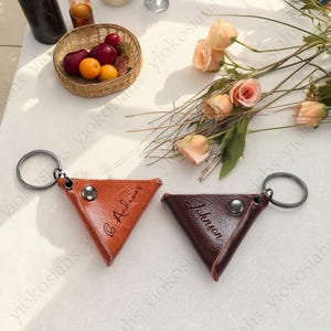 以下が含まれることがあります： 温かみのある茶色と濃いバーガンディ色の2つの革製トライアングルキーホルダー。 各キーホルダーには、シルバーの金属リングとスナップ留めが付いています。 茶色のキーホルダーには「Andreas」という名前が刻まれ、バーガンディ色のキーホルダーには「Johnson」という名前が刻まれています。