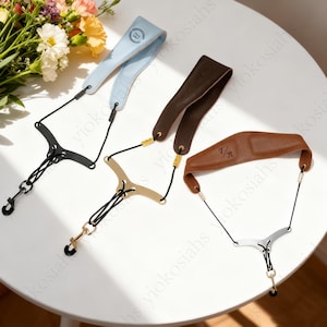 以下が含まれることがあります： 青、茶色、ベージュの革製サックスネックストラップ3本。各ストラップは異なる色とデザインで、黒いコードと金属製の金具が付いています。青いストラップには「B」の文字がエンボス加工されています。