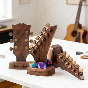 May include: Three wooden guitar headstock-shaped stands. The stands are dark brown with gold-coloured tuning pegs. One stand has the name "Sebastian" engraved on it, and another has "Jaime". Colourful guitar picks are displayed in the centre stand.