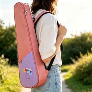 以下が含まれることがあります： 黒いバックパックストラップが付いたコーラルピンクの楽器ケース。下部のライトパープルのパネルには、カラフルな水彩画風の飛沫デザインと、黒い筆記体で「Nicole」という名前がプリントされています。ケースの側面には縦型のファスナーが付いています。