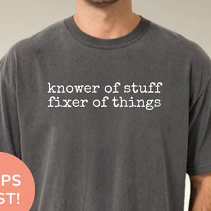 May include: A dark grey t-shirt with the text "knower of stuff fixer of things" in white. The shirt has a crew neck and short sleeves. A round peach-colored sticker with the words "SHIPS FAST!" is on the left side of the shirt.