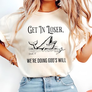 May include: Ivory t-shirt featuring the phrase "Get In Loser, We're Doing God's Will" with a whale and person graphic. The text "Jonah 18" is below the whale graphic.