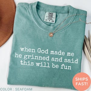 May include: Seafoam green Comfort Colors t-shirt with the text "when God made me he grinned and said this will be fun" in white. A silver necklace with a pendant rests on the shirt. The shirt is folded.
