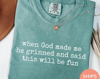 May include: Seafoam green Comfort Colors t-shirt with the text "when God made me he grinned and said this will be fun" in white. A silver necklace with a pendant rests on the shirt. The shirt is folded.