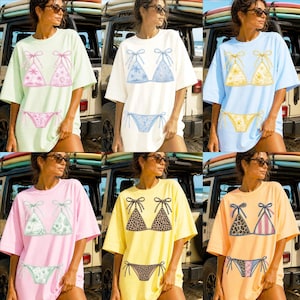 May include: A collection of oversized t-shirts in various pastel colors, each featuring a bikini top and bottom design. The designs vary, including floral, leopard print, and striped patterns. The shirts are displayed in a beach setting.
